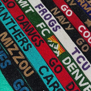 May include: A collection of long, narrow beaded straps featuring various team names and slogans including GO SUMMIT, GO DAWGS, GO FROGS, GO NAVY, GO DENVER, and GO CAROLINAS. The straps are crafted with colorful glass beads in red, green, white, black, and blue.