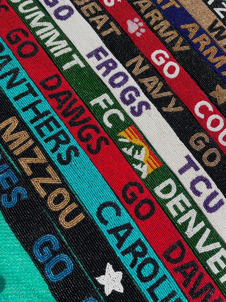 May include: A collection of long, narrow beaded straps featuring various team names and slogans including GO SUMMIT, GO DAWGS, GO FROGS, GO NAVY, GO DENVER, and GO CAROLINAS. The straps are crafted with colorful glass beads in red, green, white, black, and blue.
