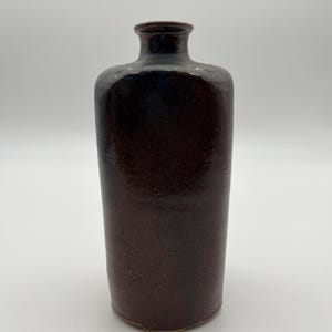 Può includere: Una bottiglia cilindrica in ceramica con una smaltatura maculata marrone scuro. Il recipiente presenta un collo stretto con un bordo leggermente svasato e una finitura liscia e lucida che sfuma in toni più profondi verso la parte superiore.