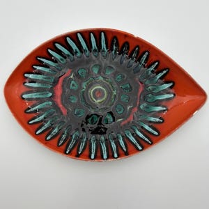 Può includere: Un piatto in ceramica a forma di occhio con un bordo arancione vivace e un centro scuro. L'interno è decorato con un motivo radiante di forme allungate color verde acqua simili a petali, che circondano un motivo circolare centrale nei toni del nero e del verde.