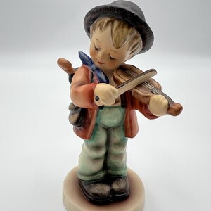 Könnte beinhalten: Eine Keramikfigur einer jungen Person mit dunklem Hut, rot-orangener Jacke und hellgrüner Hose. Die Figur spielt eine braune Geige und trägt eine kleine braune Tasche über der Schulter, alles auf einem runden Sockel montiert.