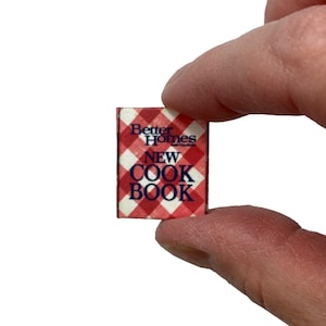 May include: A miniature book with a red and white checkered cover featuring the text Better Homes and Gardens NEW COOK BOOK in dark blue lettering. The small rectangular item has a classic kitchen-themed pattern and clear printed title text.