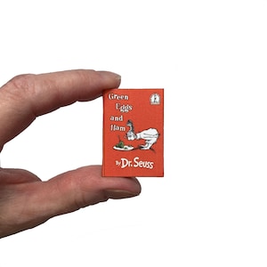 May include: A miniature book titled Green Eggs and Ham by Dr. Seuss with a bright orange cover. The front cover features a black and white illustration of a character looking at a plate of green eggs and ham. The book is small enough to be held.