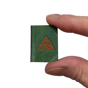 May include: A miniature green book with a textured cover featuring an embossed orange triquetra symbol centered on the front. The small rectangular volume has a vertical spine detail and is held between two fingers against a plain white background.
