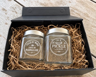 May include: Two clear glass jars with silver lids nestled in a black gift box filled with brown paper shred. The left jar features a crown icon and the text COULD BE WORSE COULD BE MARMALADE. The right jar features a pickle icon and the text IN A PICKLE...AGAIN.