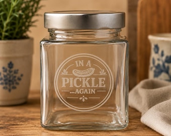 May include: A clear square glass jar with a silver-colored metal lid. The front of the jar features an etched circular design containing a graphic of a pickle and the text "IN A PICKLE ...AGAIN" with decorative lines and a small floral element.