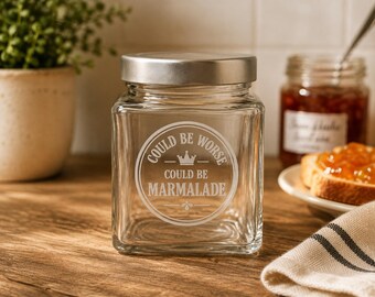 May include: A clear square glass jar with a silver-toned metal lid. The front of the jar features an etched circular design with a small crown icon and the text "COULD BE WORSE COULD BE MARMALADE" centered within the graphic lines.