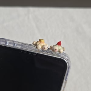 May include: Two small, cream-coloured, turtle-shaped charms attached to the top edge of a clear smartphone case. One turtle features a yellow circular accent on its shell, while the other turtle features a small, bright red pointed accent.