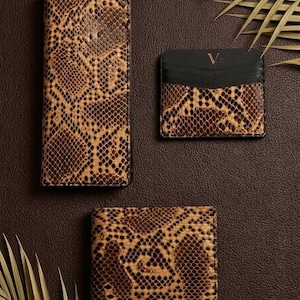 Pode incluir: Um conjunto de três acessórios com padrão de pele de cobra em tons de castanho e bege: uma carteira retangular alta, uma carteira quadrada dobrável e um porta-cartões pequeno com topo preto e a letra V gravada a dourado.