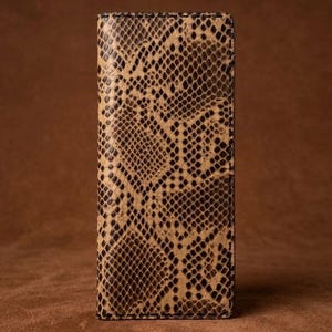 May include: A rectangular wallet featuring a textured snakeskin pattern in shades of tan, beige, and dark brown. The wallet has a vertical orientation with visible stitching along the edges, showcasing a detailed scale-like surface texture throughout.