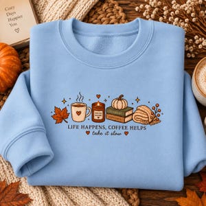 Puede incluir: Una sudadera azul claro de cuello redondo con un estampado gráfico que muestra una taza de café, una vela, dos libros apilados, una calabaza y una bufanda doblada. El texto dice: LIFE HAPPENS, COFFEE HELPS take it slow, con corazones.