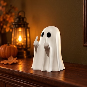 Peut inclure: Une figurine de fantôme blanche avec deux yeux ovales noirs, représentée avec les deux bras levés et les majeurs tendus. Le fantôme à la finition mate et lisse présente un aspect drapé rappelant un drap avec des plis verticaux.