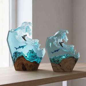 Puede incluir: Dos esculturas decorativas de resina y madera con un diseño de ola oceánica azul translúcida. Cada pieza contiene un surfista en miniatura, un tiburón y una medusa suspendidos en la resina, sobre una base de madera clara natural.