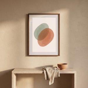 Könnte beinhalten: Ein gerahmter Kunstdruck mit zwei überlappenden, aquarellartigen Ovalen in gedecktem Salbeigrün und Terrakotta-Orange auf cremefarbenem Hintergrund, eingefasst in einen dunkelbraunen Holzrahmen mit schlichtem, klarem Rahmendesign.