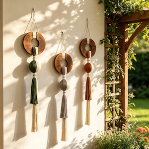 May include: Three wall hangings featuring dark brown circular wooden frames suspended by thin cords. Each piece incorporates a central sphere wrapped in yarn, with long, multi-colored tassels in shades of forest green, gray, and rust orange.