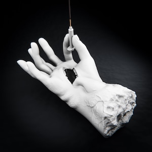 Op de afbeelding: Een wit beeldhouwwerk van een afgehakte hand met een textuur van aderen. In de handpalm bevindt zich een open mond met zichtbare tanden en de vingers houden een dun, verticaal wierookstokje vast. Het beeld staat tegen een zwarte achtergrond.
