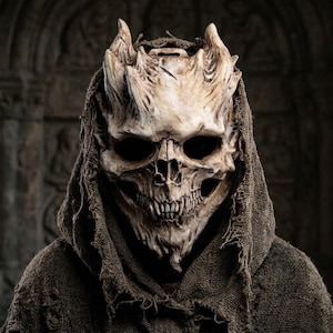 May include: A beige, weathered skull mask featuring prominent horn-like protrusions on the forehead and deep, dark eye sockets. The mask is framed by a distressed, dark brown burlap hood with frayed edges, covering the head and shoulders.