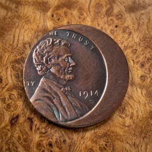 May include: A copper-coloured coin featuring a profile portrait of Abraham Lincoln facing right. The text WE TRUST is visible at the top, and the year 1914 is stamped on the right side. The coin has a distinct off-centre, crescent-shaped strike.
