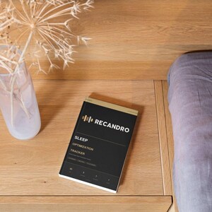 Könnte beinhalten: Ein schwarzer, rechteckiger Schlafoptimierungs-Tracker mit goldenen Akzenten und dem in Weiß gedruckten Text RECANDRO SLEEP OPTIMIZATION TRACKER. Das Gerät hat ein schlankes Design mit vier kleinen Symbolen am unteren Rand und einem goldenen Rand.