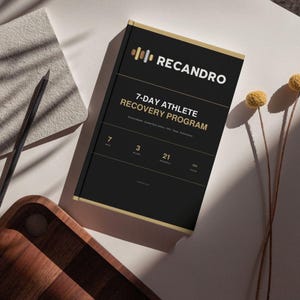 Könnte beinhalten: Ein schwarzes rechteckiges Buch mit dem Titel RECANDRO 7-DAY ATHLETE RECOVERY PROGRAM mit goldenem Text und Akzenten. Das Cover zeigt den Text 7 DAYS, 3 PHASES, 21 EXERCISES, 180 PAGES. Das Buch liegt auf einer weißen Fläche mit einem Stift.