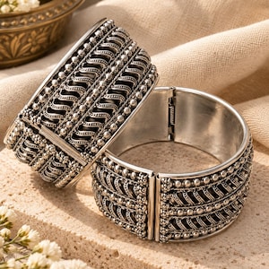 May include: Two silver-toned hinged cuff bracelets featuring intricate embossed wave patterns and rows of small spherical beads. The metal surface has a polished finish with darkened recessed areas to highlight the detailed texture of the design.