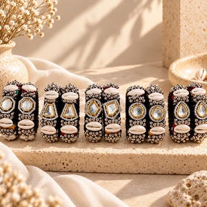 May include: Five pairs of black bangles featuring white cowrie shells, metallic silver beads, and central clear stones set in gold-toned frames. Each bangle displays a unique geometric stone shape, including hexagonal, triangular, and teardrop designs.