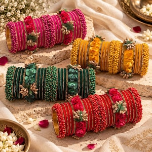 May include: Four sets of cylindrical bangle bracelets in pink, yellow, dark green, and red. Each set features a combination of smooth metallic bands, beaded textures, and decorative fabric flower accents arranged in a repeating pattern.