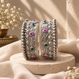 May include: Two silver-toned metal bangles featuring a textured, beaded surface with intricate geometric patterns. Each bangle is accented with small, round, embedded gemstones in alternating vibrant shades of fuchsia pink and emerald green.