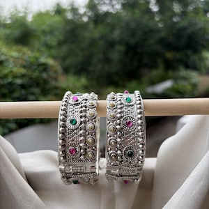 May include: Two silver-toned metal bangles featuring intricate embossed patterns, beaded edges, and small embedded circular stones in vibrant shades of pink and green. Each bangle includes a hinged opening mechanism with a secure metal clasp closure.