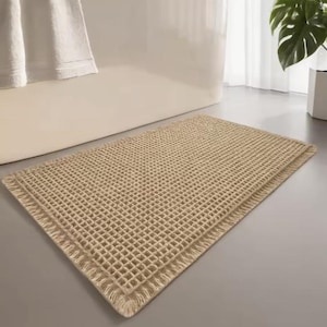 May include: A rectangular beige bath mat featuring a raised waffle-knit grid pattern across the surface. The mat has a textured, square-patterned design with a finished border edge, providing a consistent, uniform appearance throughout the piece.