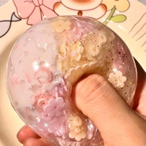 Könnte beinhalten: Ein transparentes, kreisförmiges, knautschbares sensorisches Spielzeug in Donut-Form, gefüllt mit eingebetteten rosa und weißen blumenförmigen Pailletten, irisierendem Glitzer und durchscheinendem Gel. Das Objekt wird gedrückt und zeigt seine flexible, weiche Textur.