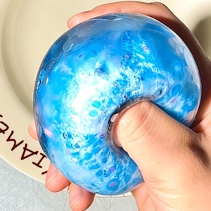 Könnte beinhalten: Ein runder, durchscheinender blauer Stressball mit einem wirbelnden Muster aus weißen und dunkelblauen Farbtönen, der kleine eingebettete Glitzerpartikel enthält. Das Objekt hat eine glatte, biegsame Textur und zeigt seine flexible Beschaffenheit.