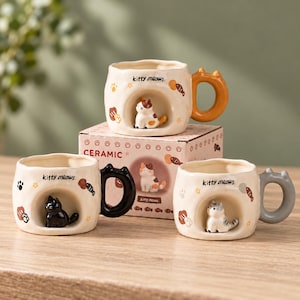 Può includere: Tre tazze in ceramica color crema con bordi irregolari, ognuna con una piccola statuetta di gatto in 3D all'interno di un incavo. Le tazze hanno manici arancioni, neri e grigi, con la scritta "kitty meows" e grafiche di pesci e impronte.