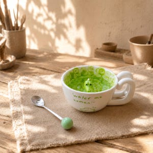 Puede incluir: Una taza de cerámica blanca con asa de doble bucle y un interior verde que presenta una pequeña cabeza de cocodrilo esculpida. El exterior tiene el texto "water me human" en verde y un dibujo de un cocodrilo, junto a una cuchara plateada.