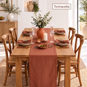 Op de afbeelding: Een rechthoekige tafelloper van linnen in de kleur terracotta, gecentreerd op een eettafel van licht hout. De tafel is gedekt met witte borden, terracotta servetten, wijnglazen, goudkleurig bestek en een centrale vaas met een kaars.