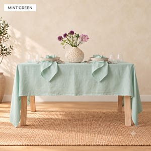 May include: A rectangular mint green tablecloth draped over a wooden table. The table is set with two place settings featuring mint green napkins, plates, and clear glasses. A textured white vase with purple and pink flowers sits in the center.