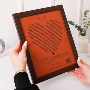 May include: A dark brown wooden frame containing a burnt orange print with the song lyrics to All of Me by John Legend arranged in a heart shape. The print includes musical notes, a QR code, and the names Christy & Daniel with a date.