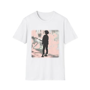 May include: A white short-sleeve t-shirt featuring a central graphic print of a silhouette of a person with messy hair holding a guitar, set against a soft, blurred background of pale pink and grey tones with a rectangular border.