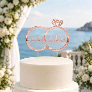 May include: A rose gold cake topper featuring two interlocking rings, one topped with a diamond shape, and the names Isabella & Noah in script font. The topper is supported by two clear acrylic stakes and sits atop a smooth white cake.
