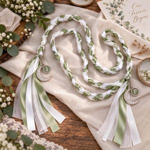 May include: A braided cord made of white and sage green satin ribbons, accented with white lace trim. Each end features a silver-toned crescent moon charm with clear rhinestones and a circular pendant displaying the letters J and P respectively.
