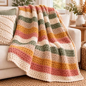May include: A crocheted throw blanket featuring horizontal stripes in alternating colours of cream, sage green, dusty pink, and mustard yellow. The blanket has a textured stitch pattern and a simple border, draped over the edge of a light-coloured sofa.