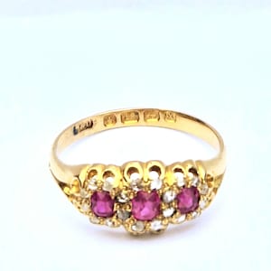 May include: A gold ring featuring three central cushion-cut red rubies surrounded by small clear stones. The band displays visible hallmarks on the interior surface, including a clear stamp of 375, indicating the metal composition of the jewellery piece.