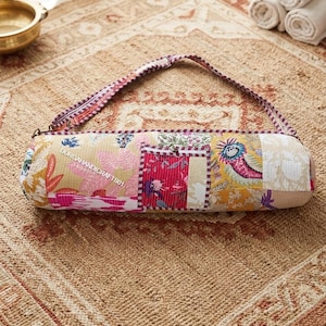 May include: A cylindrical yoga mat bag featuring a patchwork design with various floral and abstract patterns in shades of yellow, pink, red, and white. The bag includes a long adjustable shoulder strap and a small square pocket with the text DURGAHANDICRAFTS01.