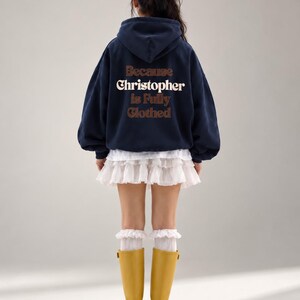 Könnte beinhalten: Ein marineblauer Kapuzenpullover mit dem Schriftzug Because Christopher is Fully Clothed in braunen und weißen Buchstaben auf der Rückseite. Das Kleidungsstück wird mit einem weißen Minirock mit Rüschen, weißen Rüschensocken und gelben Stiefeln kombiniert.
