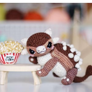 Peut inclure: Un jouet Ankylosaurus au crochet nommé Bruno en fil marron et crème, avec des pointes blanches sur le dos, de grands yeux noirs et des joues roses, placé à côté d'un petit bol de pop-corn avec le texte POP CORN sur un gobelet rayé rouge et blanc.