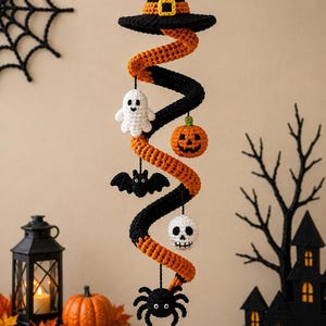 Könnte beinhalten: Eine hängende Häkelspirale, die oben mit einem schwarzen Hexenhut mit gelber Schnalle versehen ist. Die orange-schwarze Spirale ist mit kleinen gehäkelten Figuren geschmückt, darunter ein weißes Gespenst, ein orangefarbener Kürbis, eine schwarze Fledermaus, ein weißer Totenkopf und eine schwarze Spinne.
