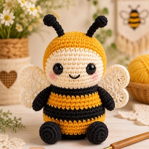 Könnte beinhalten: Ein gehäkeltes Bienen-Stofftier mit rundem Körper, horizontalen gelben und schwarzen Streifen, einem cremefarbenen Gesicht, großen schwarzen Sicherheitsaugen, rosa Wangen, zwei schwarzen Fühlern und zarten weißen Spitzenflügeln am Rücken.