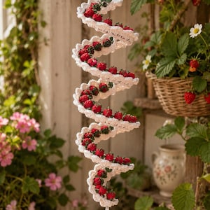 Könnte beinhalten: Eine weiße, gehäkelte Spiral-Hängedekoration mit zahlreichen kleinen roten Erdbeeren mit grünen Blättern entlang der Windungen, die an der untersten Spitze der Spiralstruktur mit einer einzelnen, größeren roten Erdbeere endet.