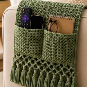 Könnte beinhalten: Ein olivgrüner, gehäkelter Armlehnen-Organizer mit zwei tiefen Taschen, einer dekorativen Bordüre im Gittermuster und einer Reihe hängender Quasten. Die Taschen enthalten eine schwarze Fernbedienung, ein Smartphone, ein Notizbuch und eine Brille.