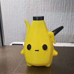 May include: A yellow, low-poly, 3D-printed banana-shaped figurine with a black cap on top. The figure features two round black eyes and a small horizontal black line for a mouth, with small protruding arms on the sides and a curved stem-like element.